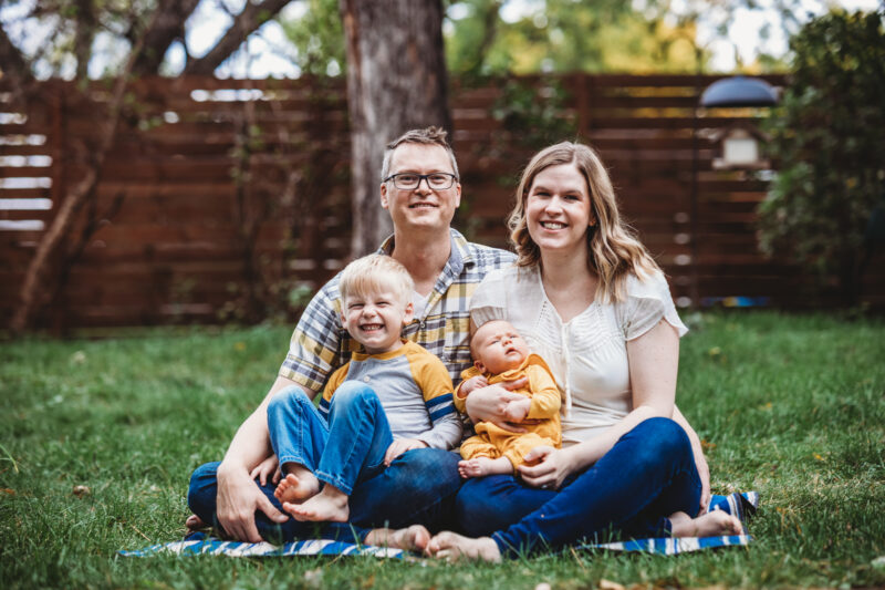A mother and father sitting in the grass holding their toddler and baby on their lap.