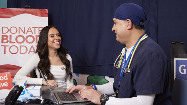 NYBC doctor discussing donation process with blood donor. Donate Blood Today sign is in the background.
